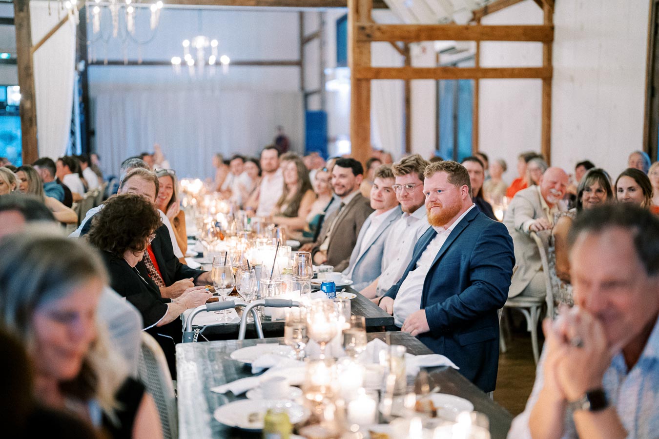 A large group of people seated at elegantly decorated tables during a formal event, with soft candlelight and chandeliers creating a warm ambiance in a rustic venue.