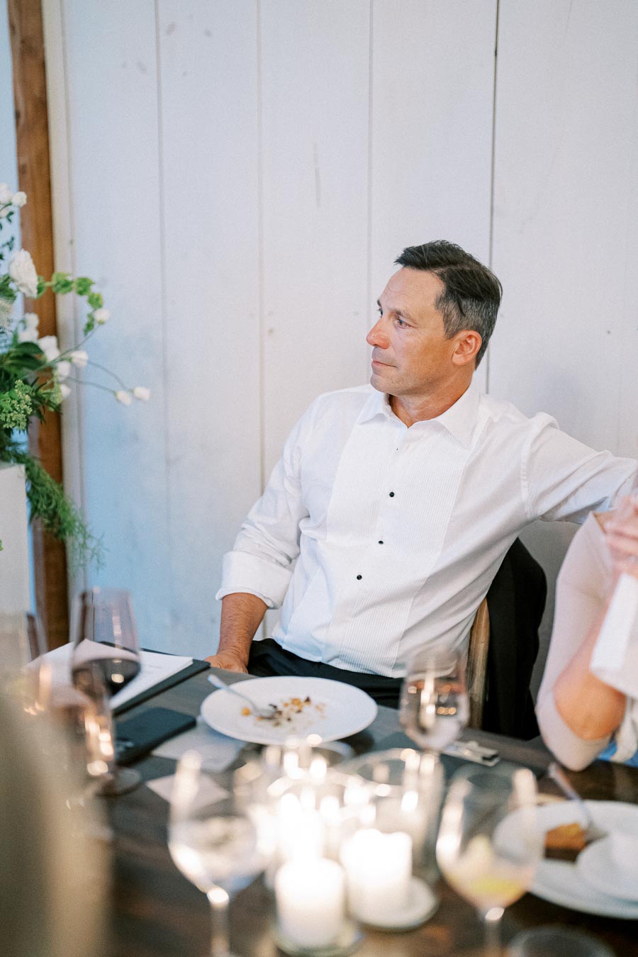 A man in a white dress shirt sitting at an elegantly set dining table, surrounded by candles and glassware, with a partially eaten meal on a plate in front of him.