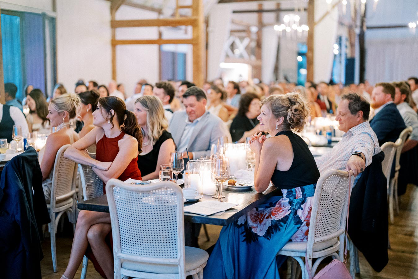 Guests seated at tables during an elegant indoor event, attentively listening, surrounded by soft candlelight and sophisticated decor.