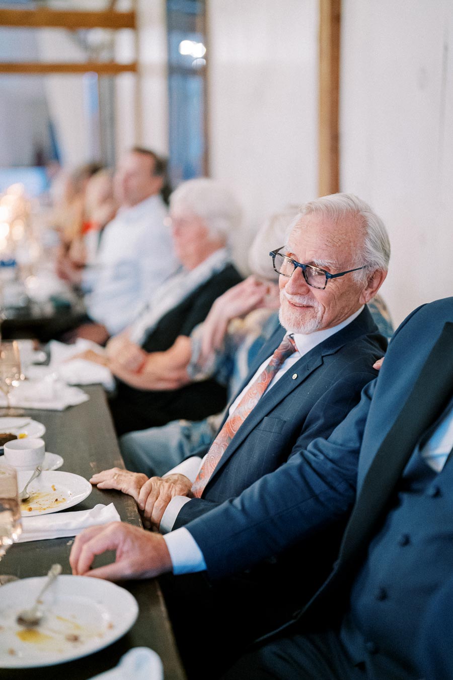 Senior man in formal attire smiling at a dinner event, surrounded by other guests seated at a long table, creating a cheerful and elegant atmosphere.
