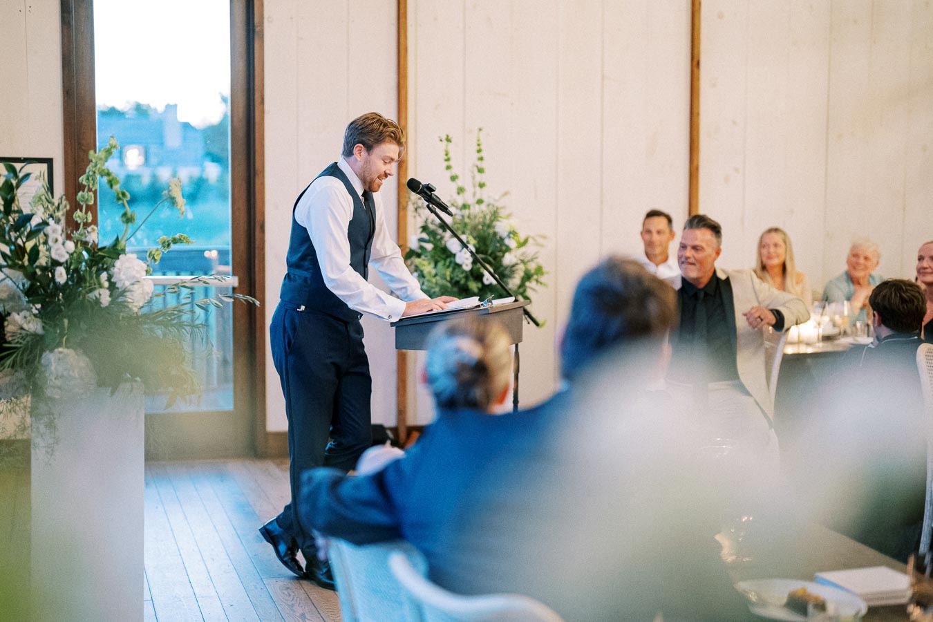 A man in formal attire, standing at a podium with a microphone, delivers a speech at an indoor event with attendees seated around tables, and floral arrangements in the background.
