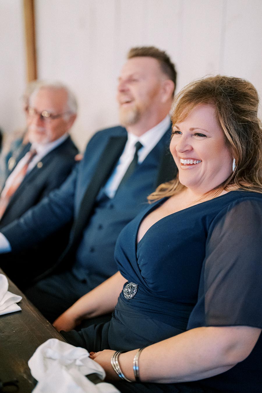 Three people seated at a formal event, smiling and dressed in elegant attire, including a woman in a navy dress and a man in a suit.