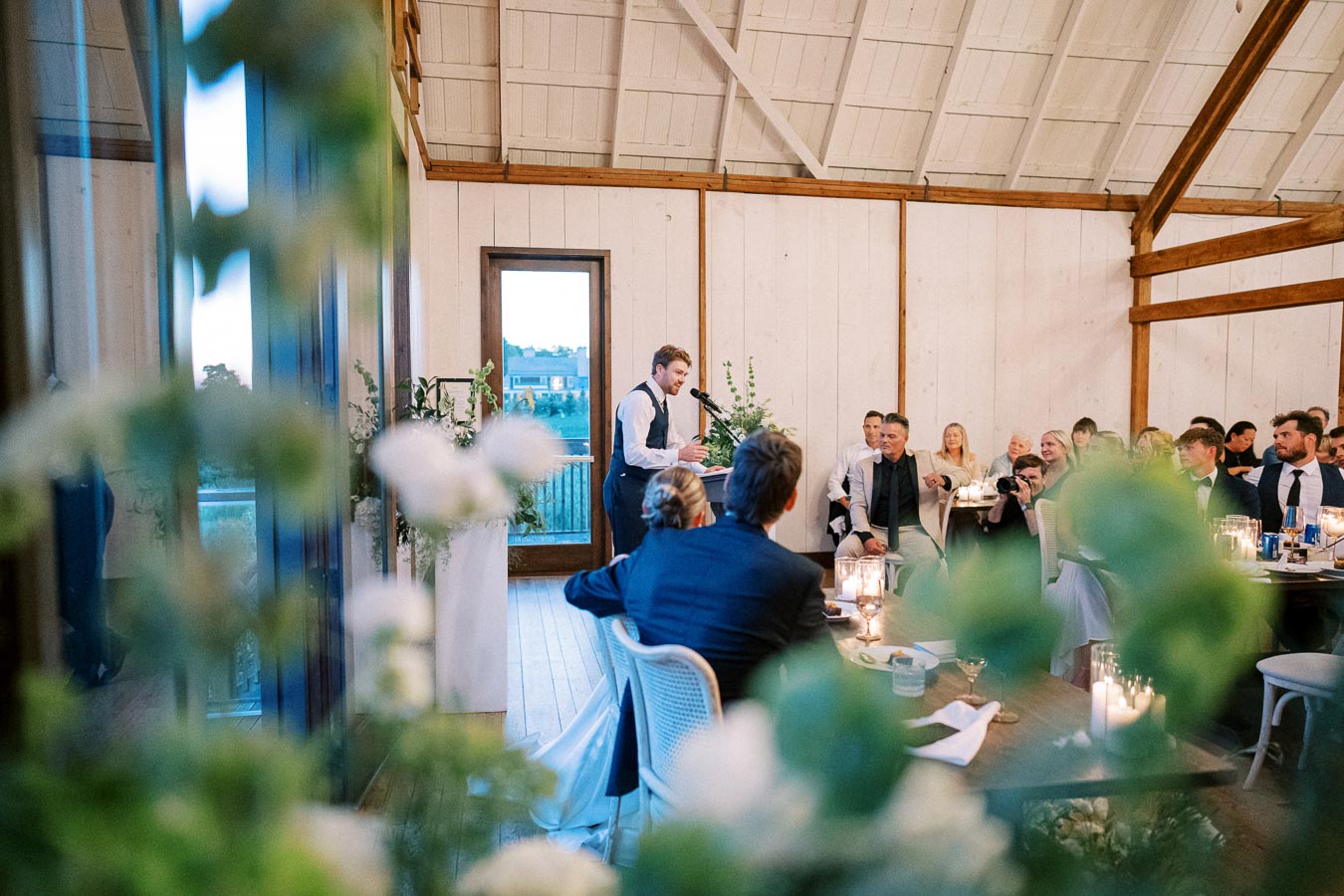 Wedding reception speech at a rustic venue with a wooden interior, a speaker at a microphone, guests seated at candle-lit tables, and floral decorations creating a warm, festive atmosphere.