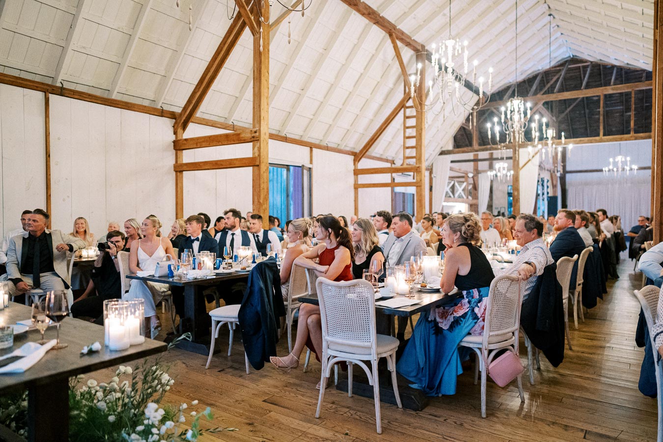 Guests seated at elegantly decorated tables inside a rustic barn venue during a wedding reception, with ambient lighting from chandeliers and candlelit centerpieces.