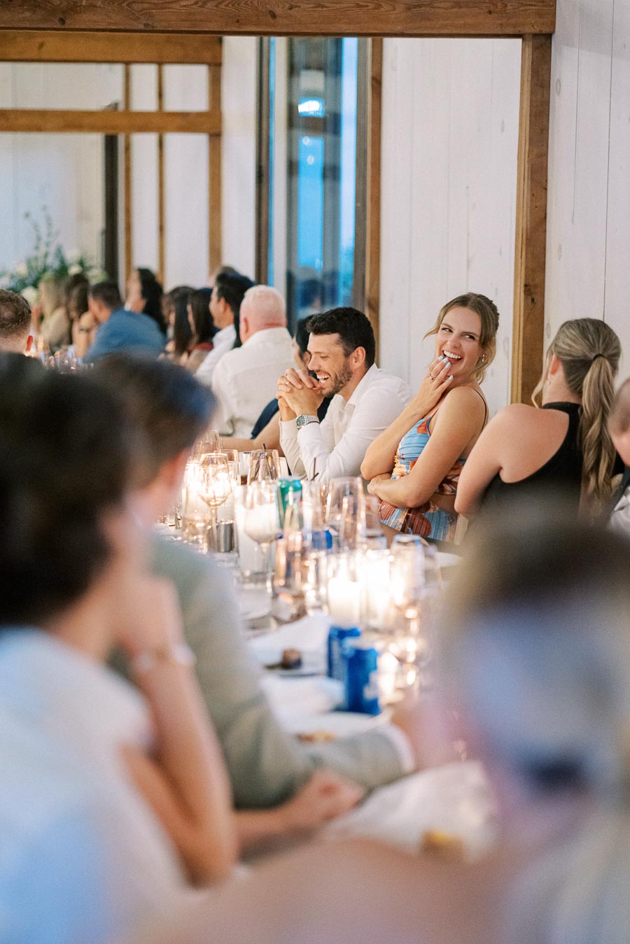Guests laughing and enjoying a lively reception dinner at an elegantly decorated wedding venue, featuring candlelit tables and wooden rustic decor.