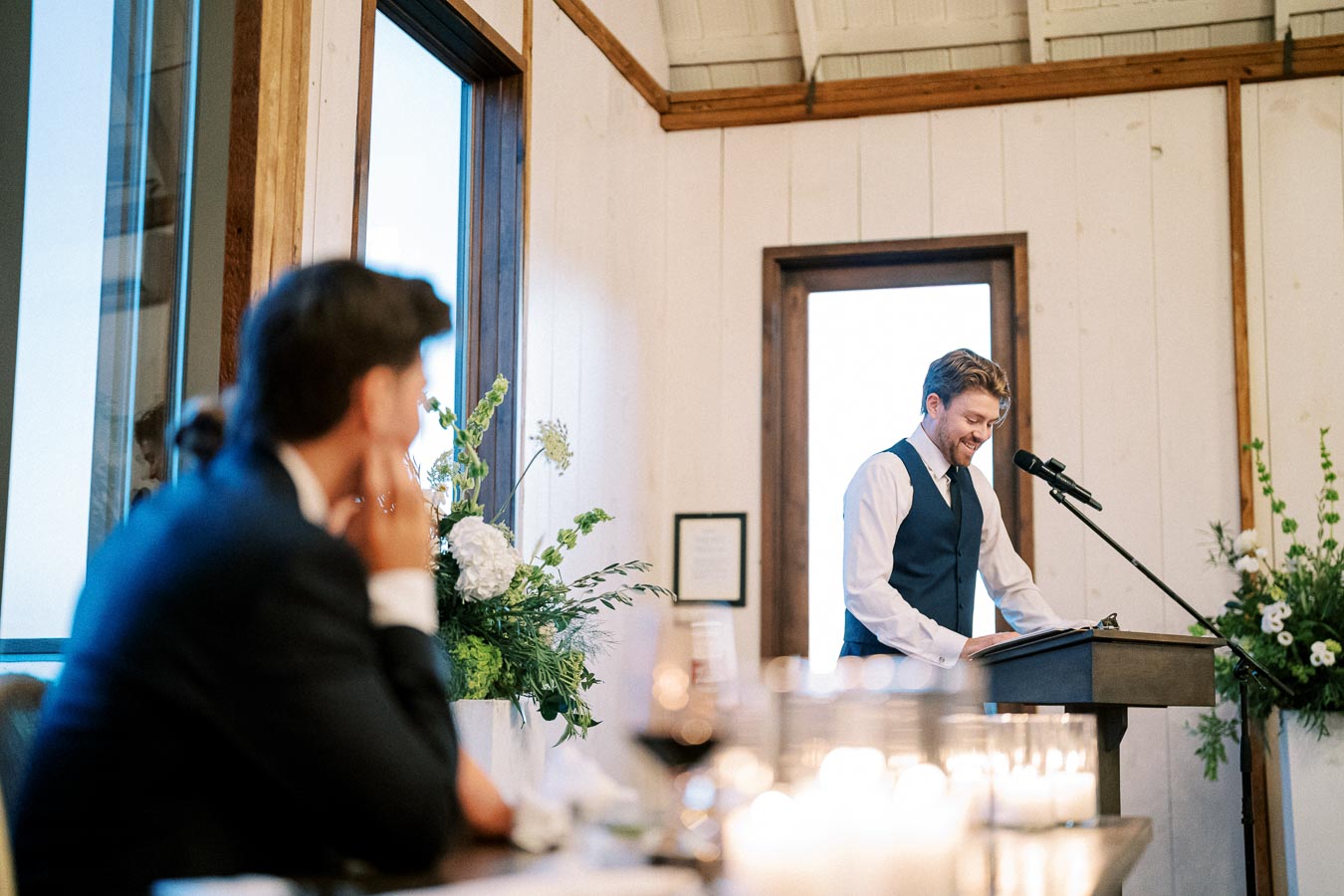 Man in a suit giving a speech at a wedding reception, standing at a podium with candles and floral arrangements in the foreground, creating a warm and elegant ambiance.
