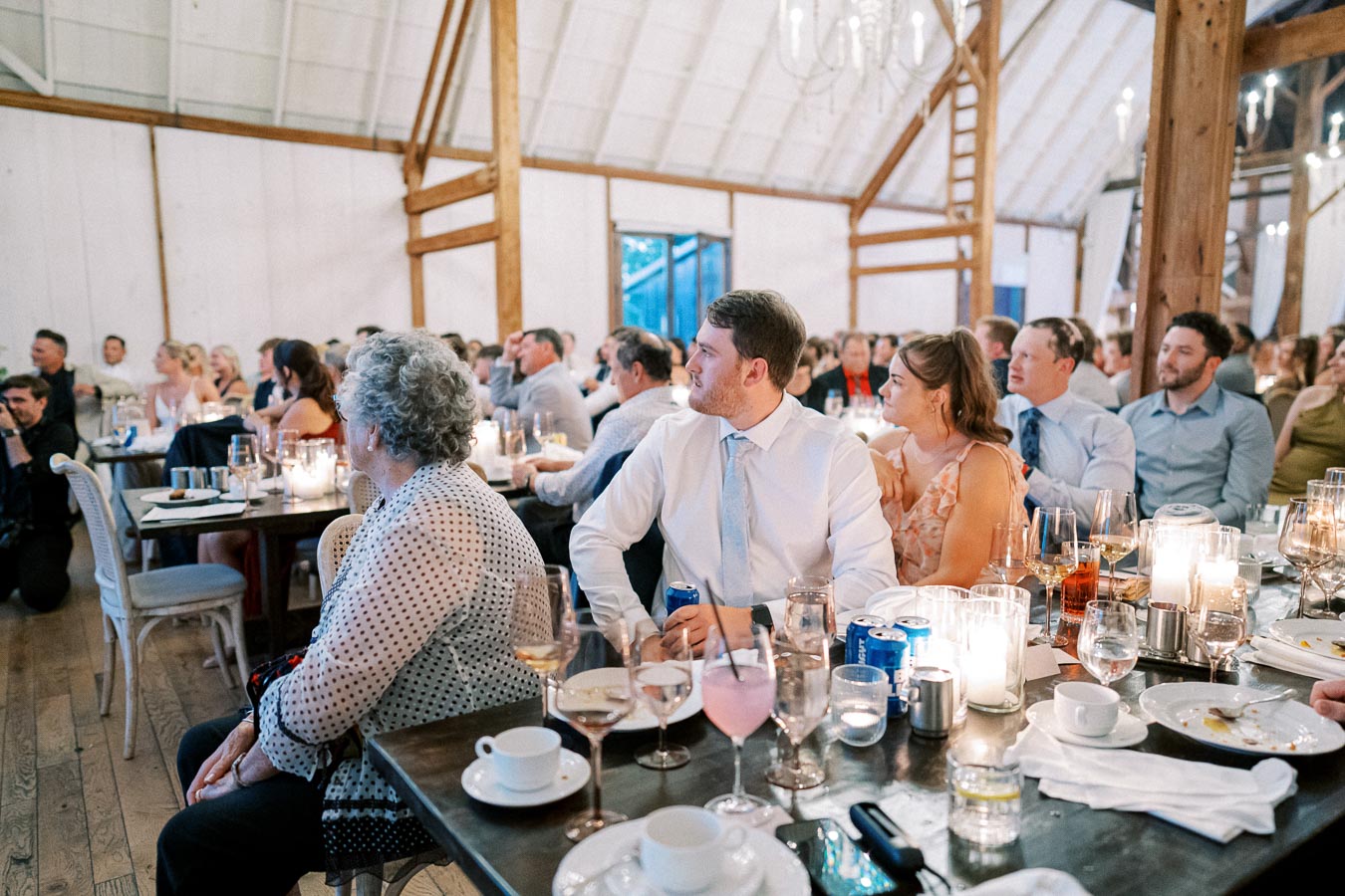 Guests seated at a rustic wedding reception in a barn venue with wooden beams, enjoying a formal dinner. Tables are set with candles, drinks, and elegantly arranged dinnerware, creating a warm and inviting atmosphere.