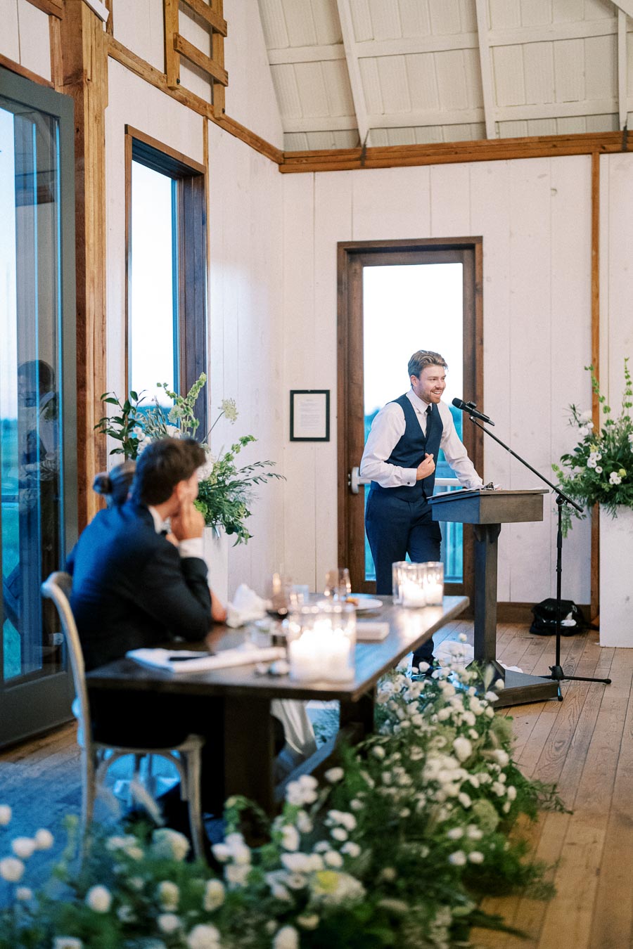 Groomsman giving a heartfelt speech at a wedding reception, with guests seated at a beautifully decorated table adorned with candles and floral arrangements, in a rustic venue with wooden accents.