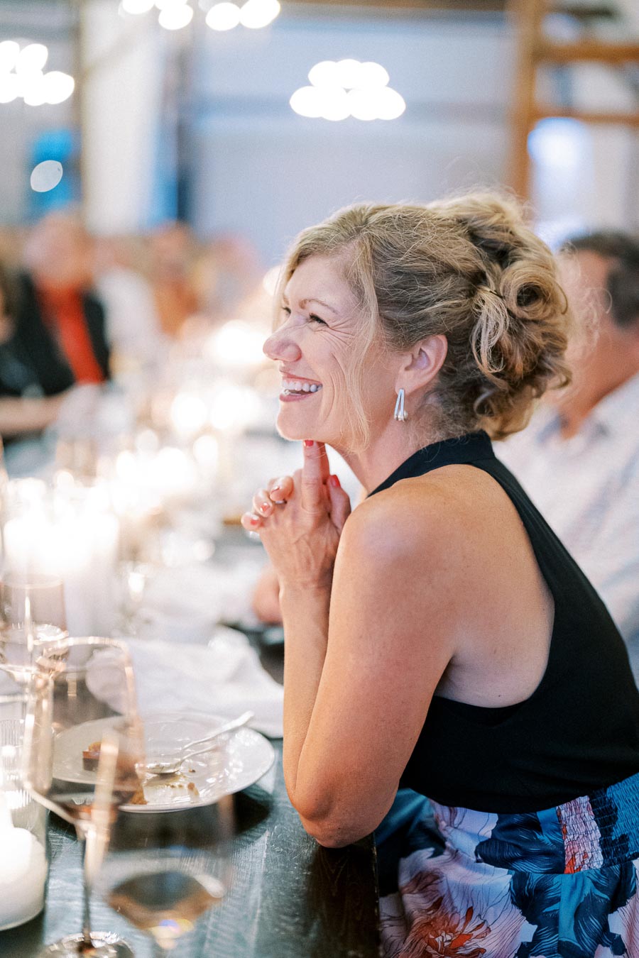 Smiling woman enjoying dinner at a warmly lit restaurant table, surrounded by soft lighting and blurred guests in the background.