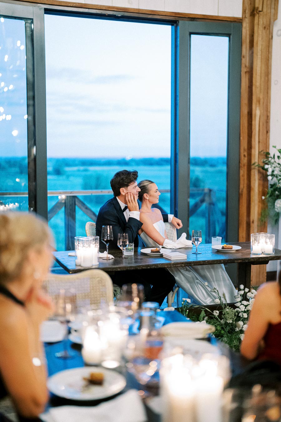 Elegant wedding reception with a couple seated at a table, enjoying the view through large windows, surrounded by candlelight and floral arrangements.