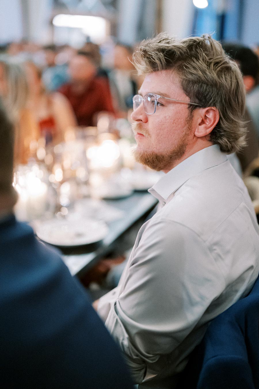 Man wearing glasses and a white shirt seated at a formal event with blurred guests in the background.