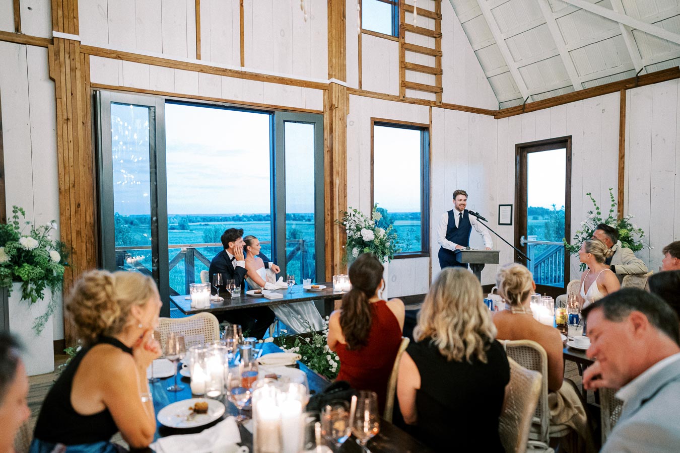 Wedding reception speech at rustic venue with guests seated at candlelit tables, overlooking scenic outdoor view through large windows.