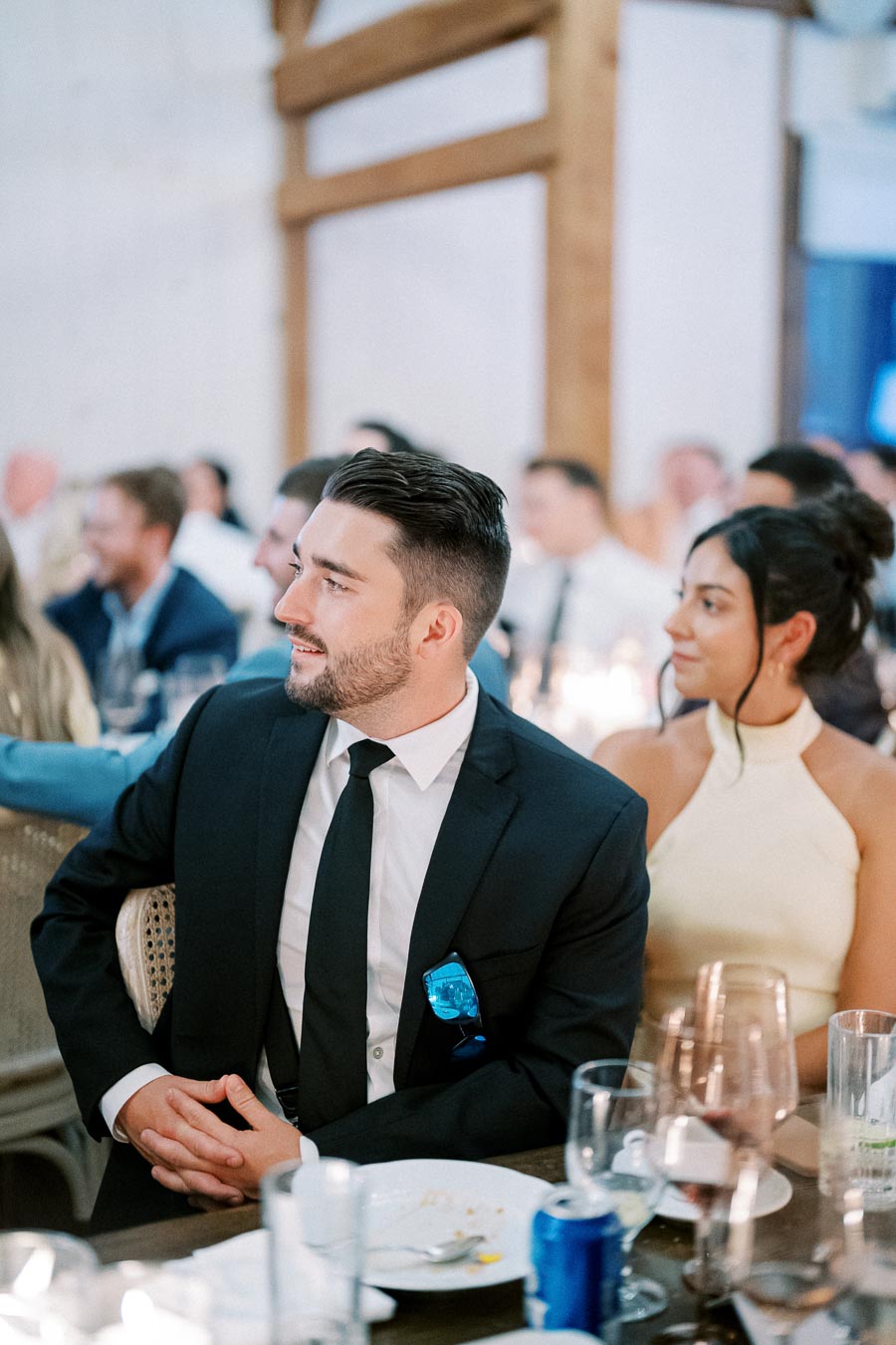 A well-dressed man in a black suit and tie sits attentively at a formal event next to a woman in a cream halter dress, with a blurred background of other attendees at a banquet.