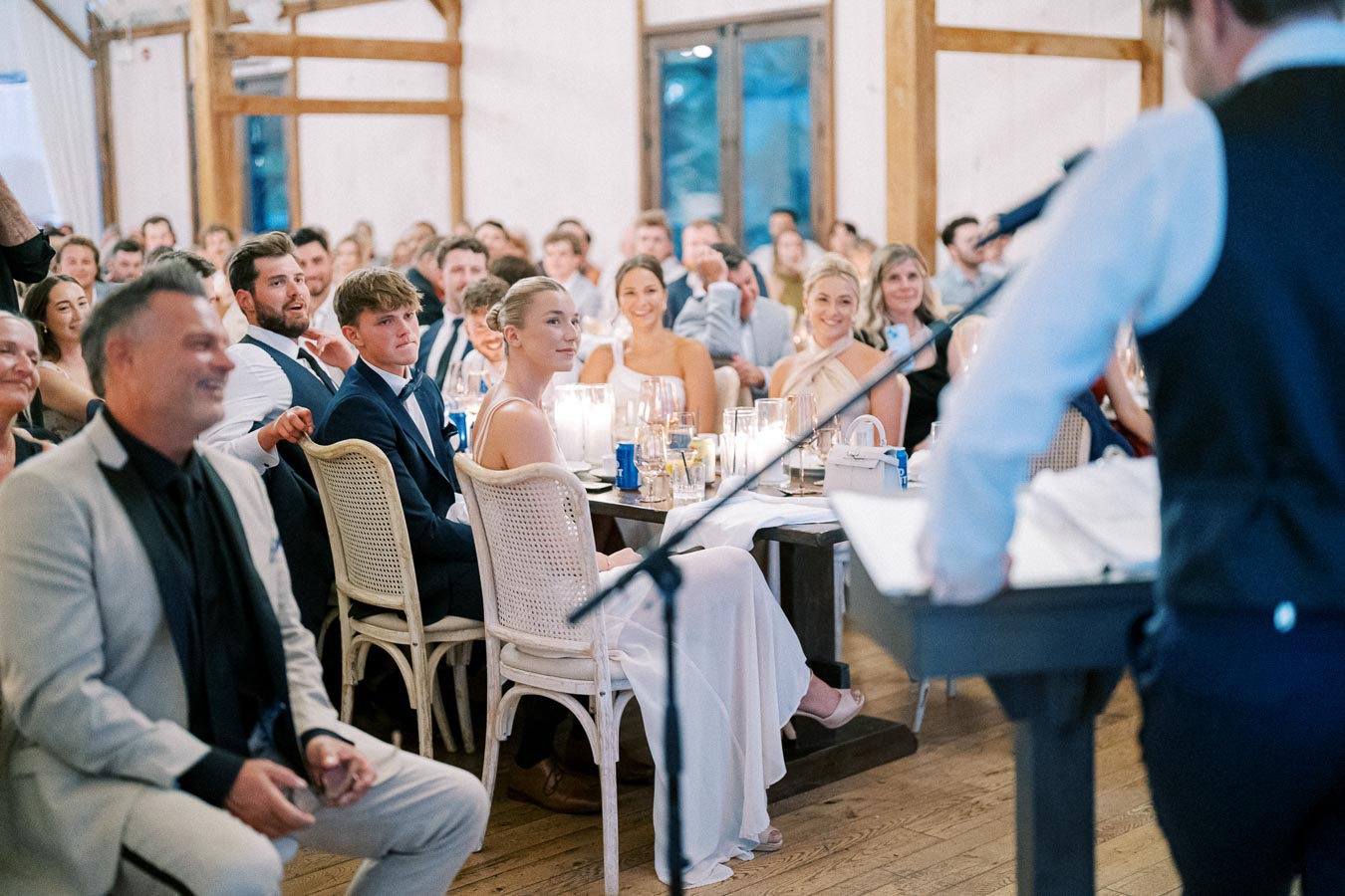 Wedding reception scene with a crowd attentively listening to a speaker, seated guests in elegant attire inside a rustic, wooden venue.