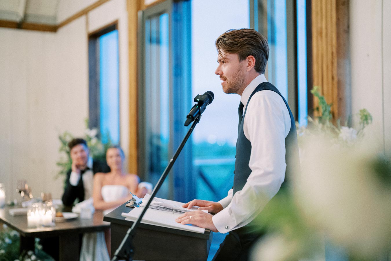Best man delivering a heartfelt speech at a wedding reception, with the bride and groom smiling in the background, set in a beautifully decorated venue.