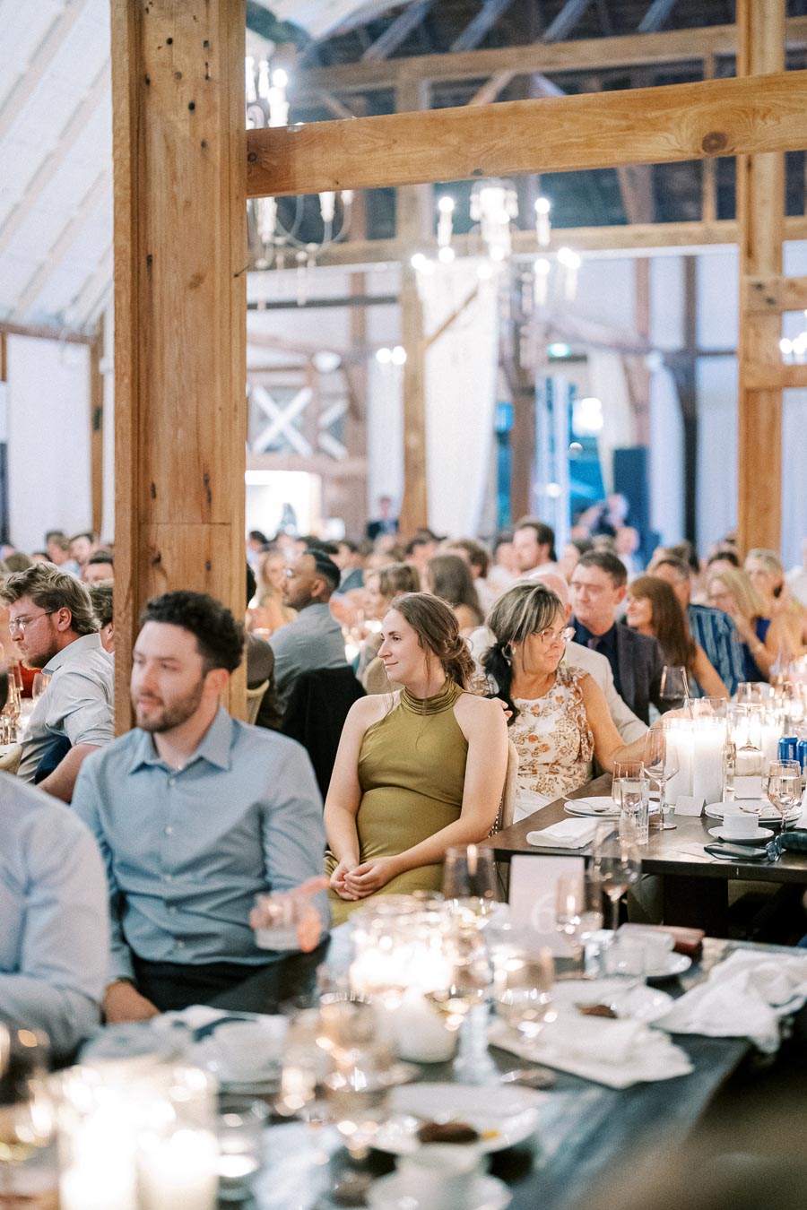 Guests seated at a beautifully decorated wedding reception inside a rustic barn, with tables adorned with candles and dinnerware, creating a warm and cozy atmosphere.