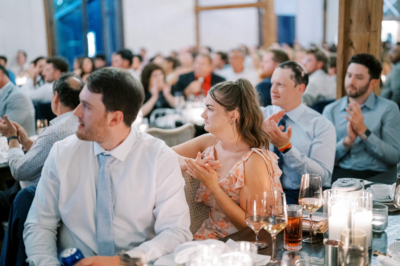 Audience at formal event clapping, featuring elegantly dressed attendees at dining tables, with visible candles and drinks, suggesting an upscale gathering or celebration.