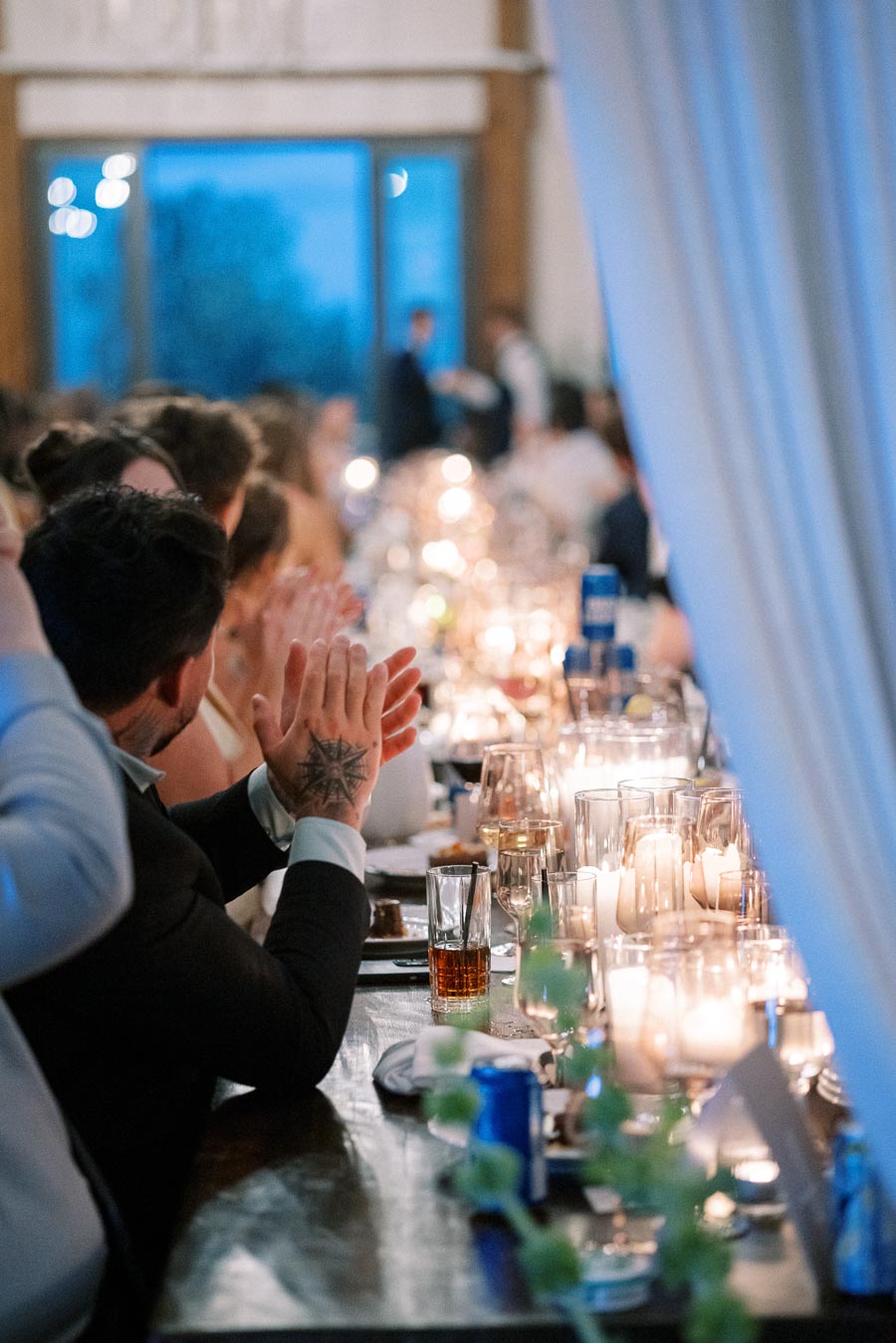 A warmly lit banquet hall with guests seated at a long dining table, elegantly set with glasses and candles, creating an inviting ambiance, as attendees applaud during a formal event.