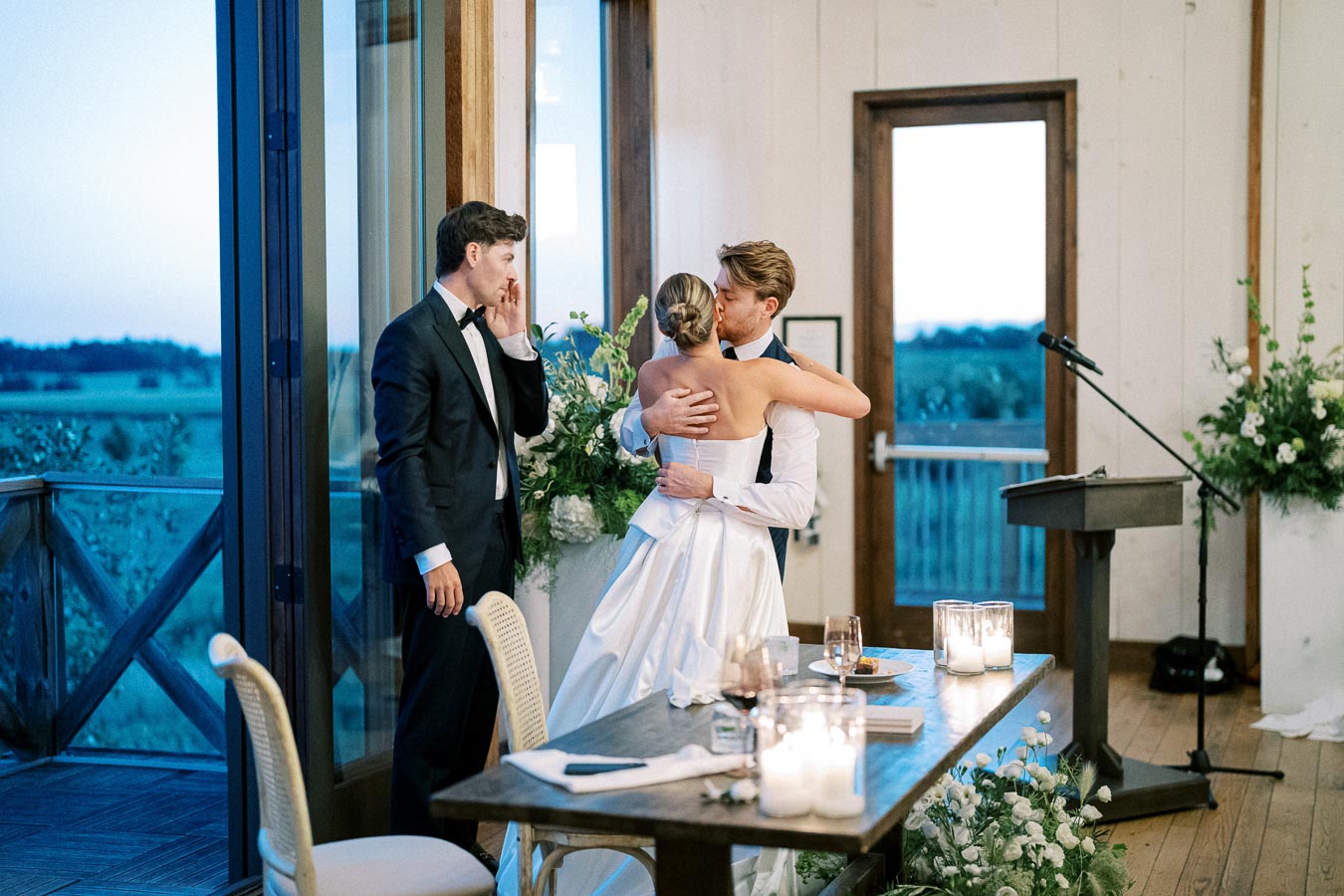 A bride in a white gown and a groom in formal attire share a loving embrace during their wedding reception, with a groomsman in the background. The indoor setting features elegant decor with candles, flowers, and a scenic outdoor view through large windows.