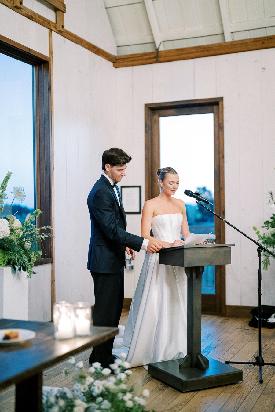 A bride in a white wedding dress and a groom in a black suit stand at a podium, delivering a speech during their wedding ceremony in a bright, elegant room with large windows and floral arrangements.