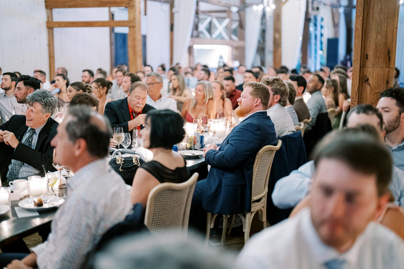 A lively indoor formal dinner event, featuring guests dressed in suits and evening attire, seated around elegantly set tables with sparkling glassware and candlelight.