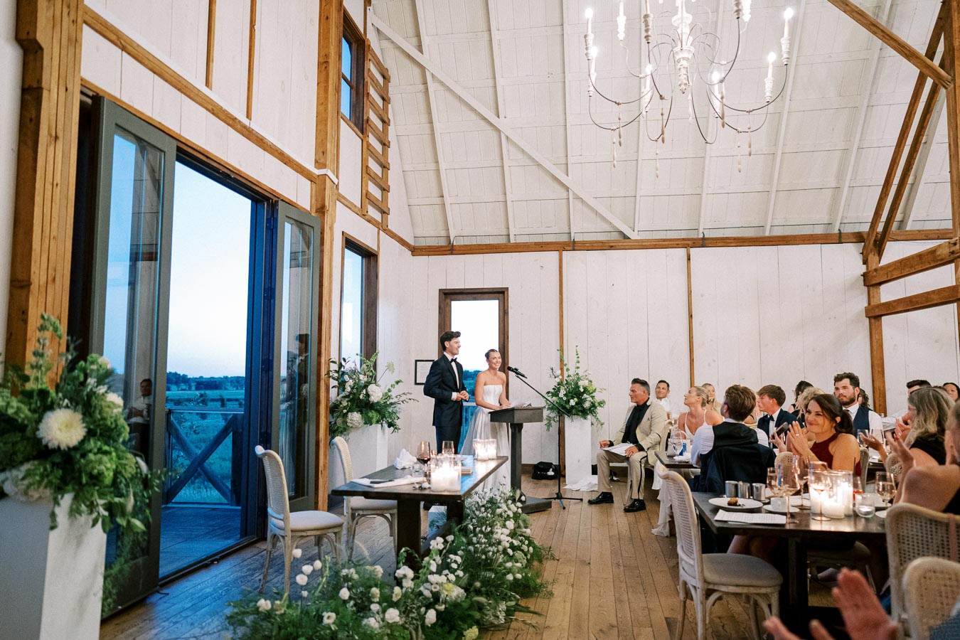 Elegant rustic wedding reception in a beautifully decorated barn venue, featuring guests seated at candlelit tables, and a couple delivering a speech at a modern podium surrounded by greenery.