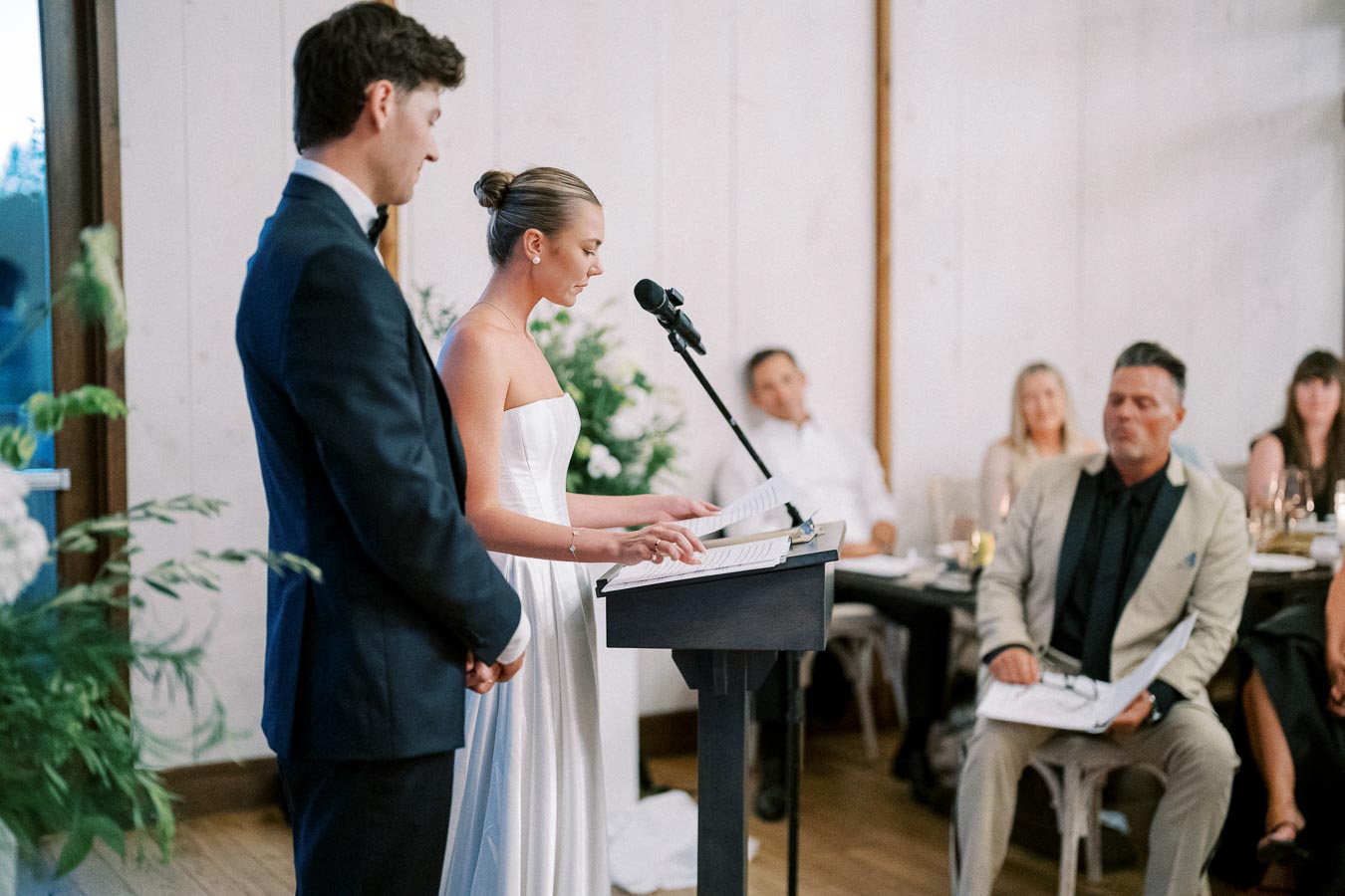A couple giving a heartfelt speech at a wedding reception, with attentive guests seated in the background, in a beautifully decorated venue.