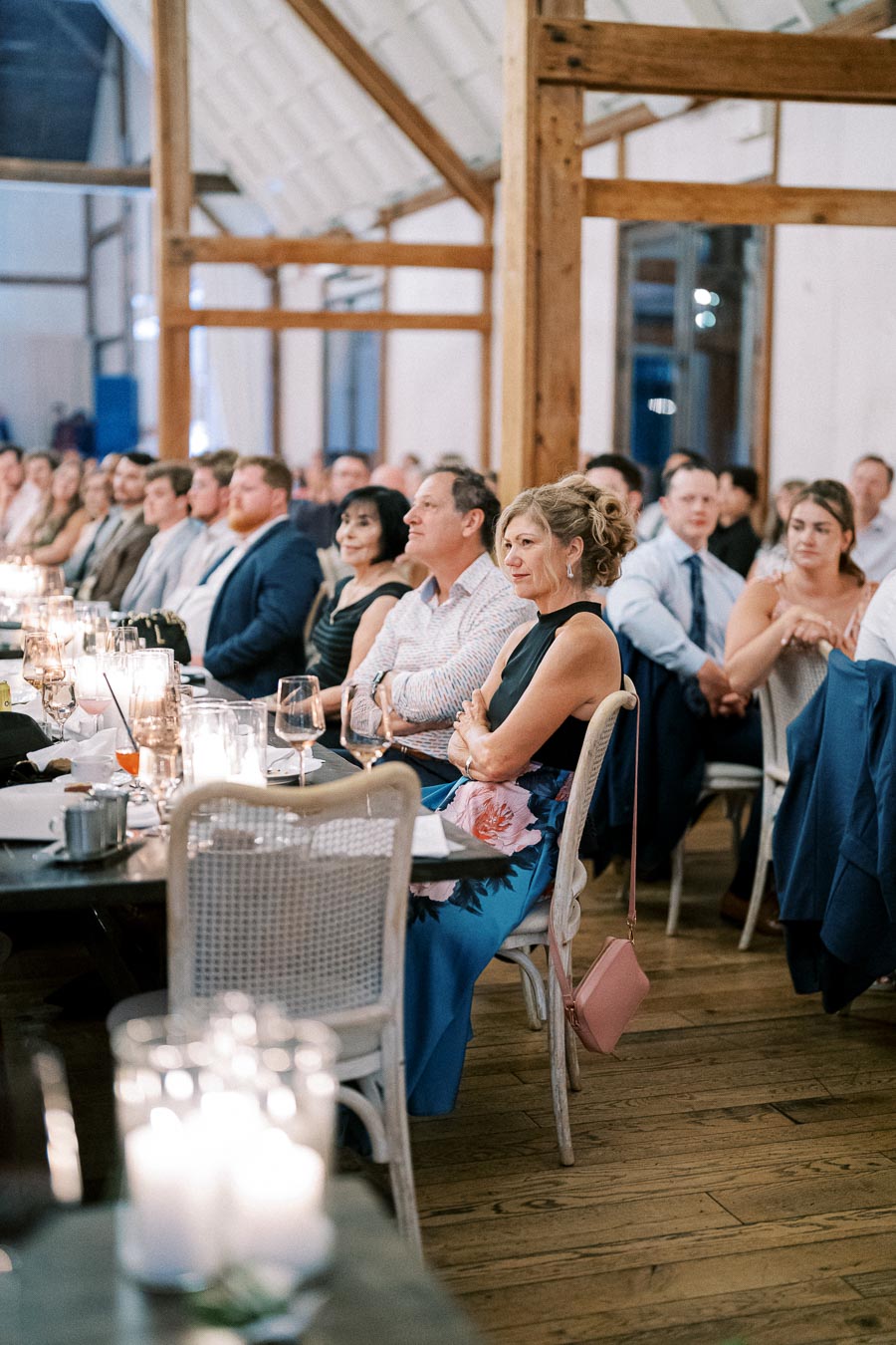 A group of elegantly dressed guests seated at a rustic wedding reception in a wooden barn venue, with soft candlelight on the tables creating a warm and intimate atmosphere.