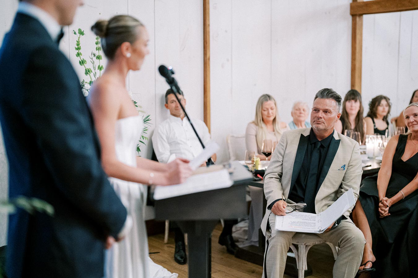 A bride in a white dress delivers a speech at a wedding reception, while a man in a light suit attentively listens with a script in his hand. Guests are seated around a table decorated with candles and wine glasses, creating an elegant atmosphere.