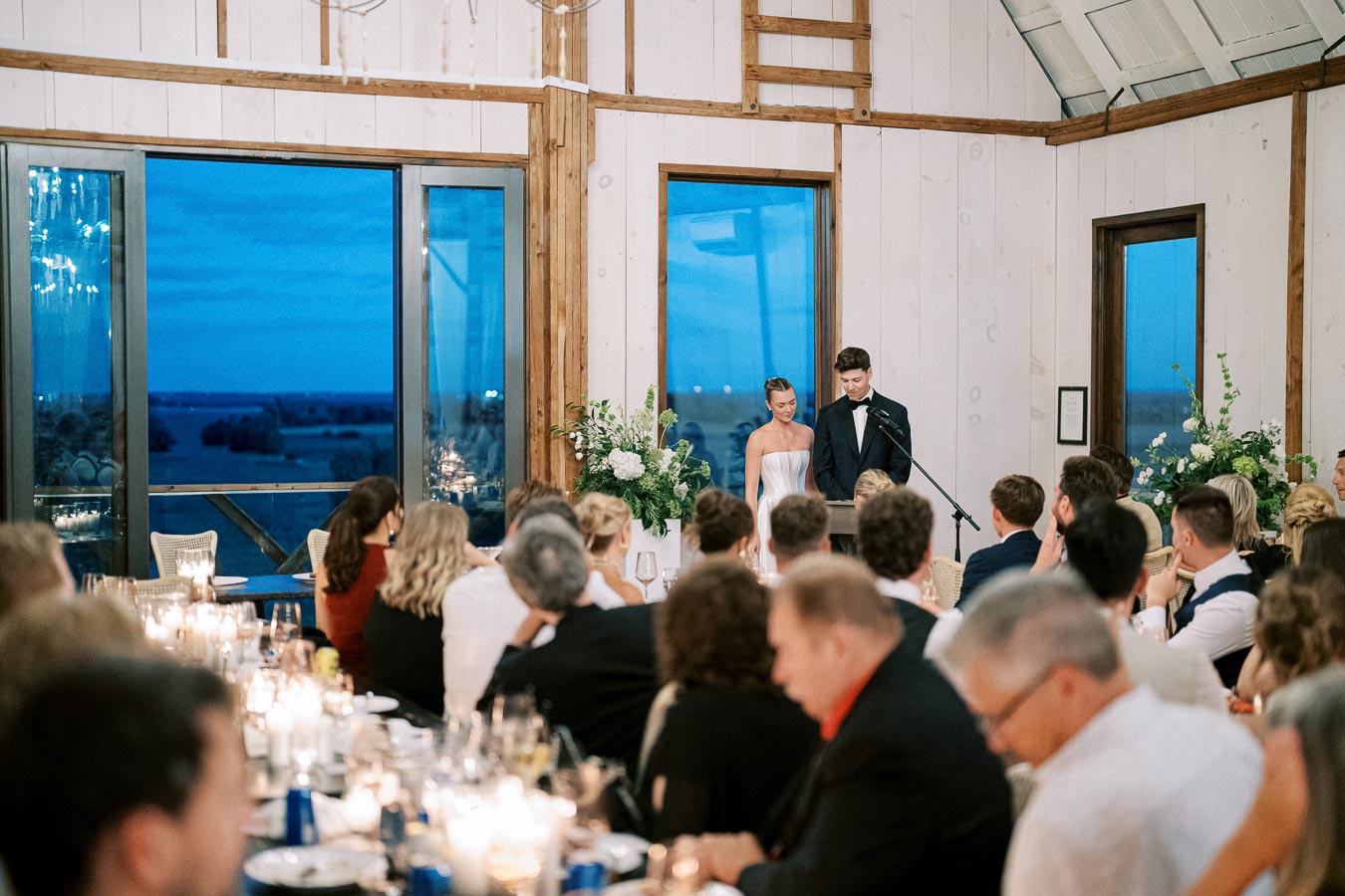 Elegant wedding reception with a couple giving a speech at the head table, surrounded by attentive guests, in a beautifully decorated venue with large windows showcasing a scenic evening view.