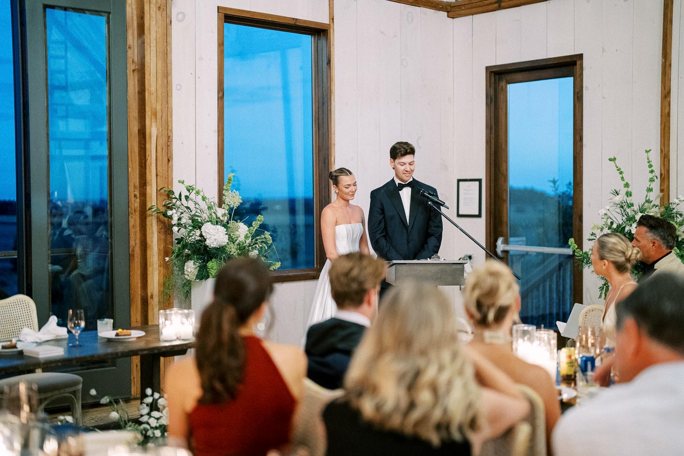 A bride and groom giving a speech at their wedding reception in an elegantly decorated room with floral arrangements and seated guests listening attentively.