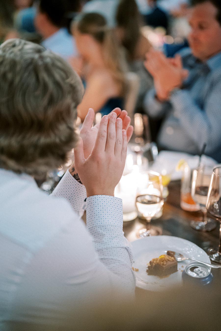 A person in a white shirt clapping at a dinner event, with a partially eaten dessert and beverages on the table.