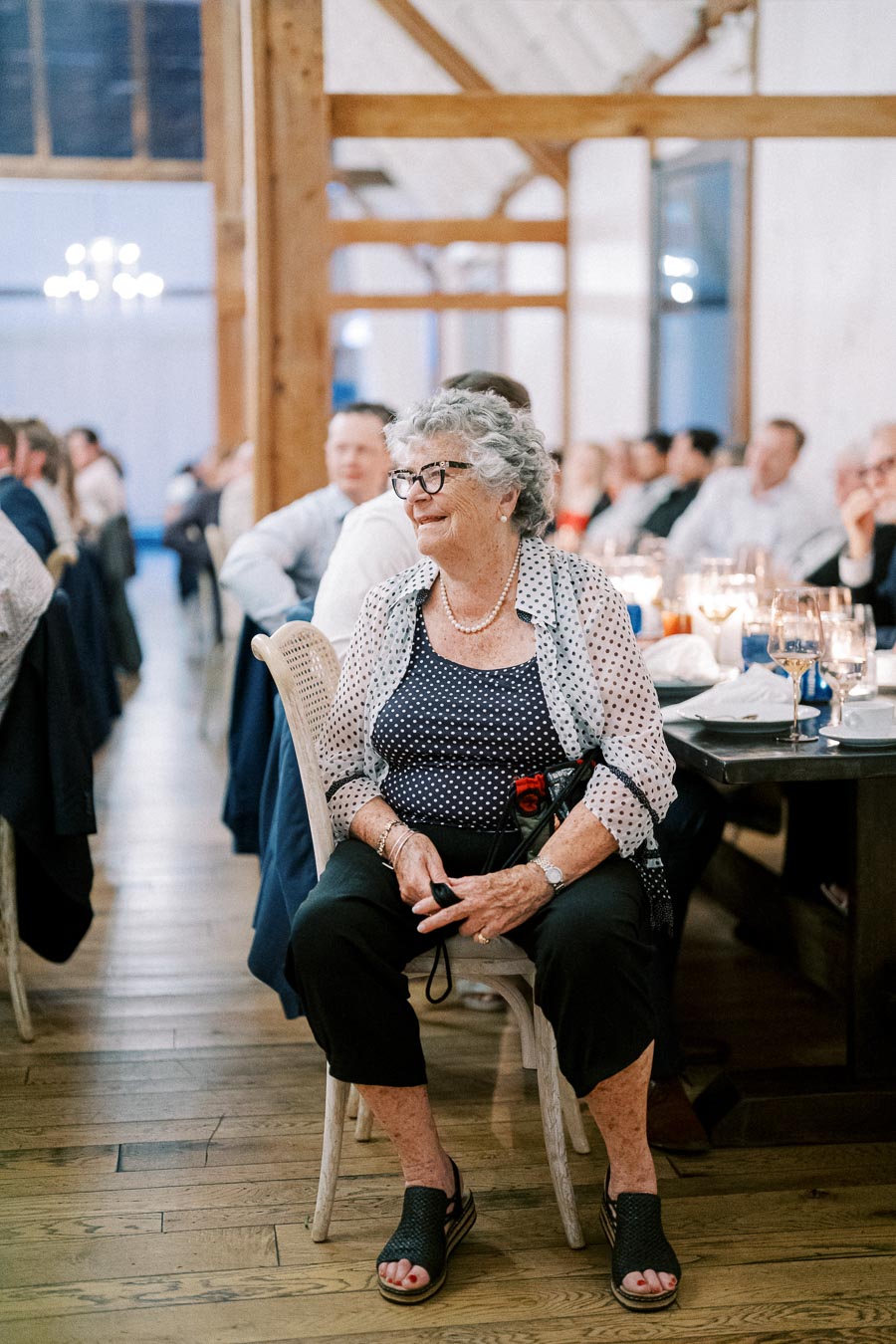 Elderly woman sitting and smiling at a formal event, surrounded by guests at tables in a warmly lit banquet hall.