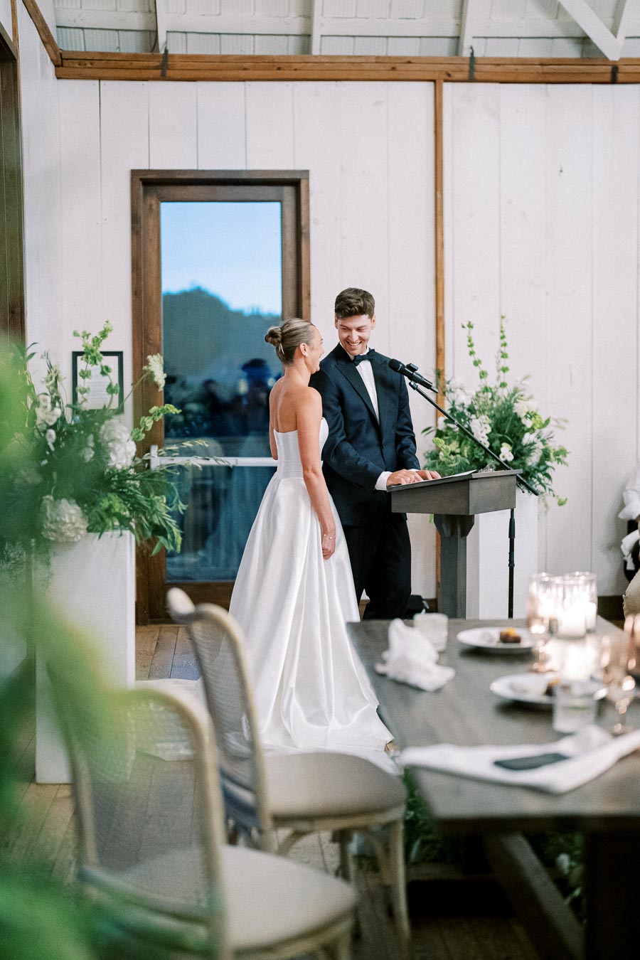 A bride in an elegant white dress and a groom in a black tuxedo share a joyful moment while speaking at a wedding reception podium, surrounded by floral decorations and candlelit tables.