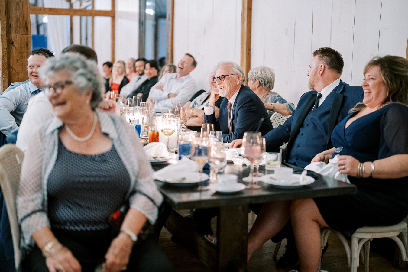A group of people sitting at a long table during an indoor event, laughing and enjoying a lively moment. The room is warmly lit, and the table is adorned with glasses, dishes, and candles, contributing to a cheerful and festive atmosphere.