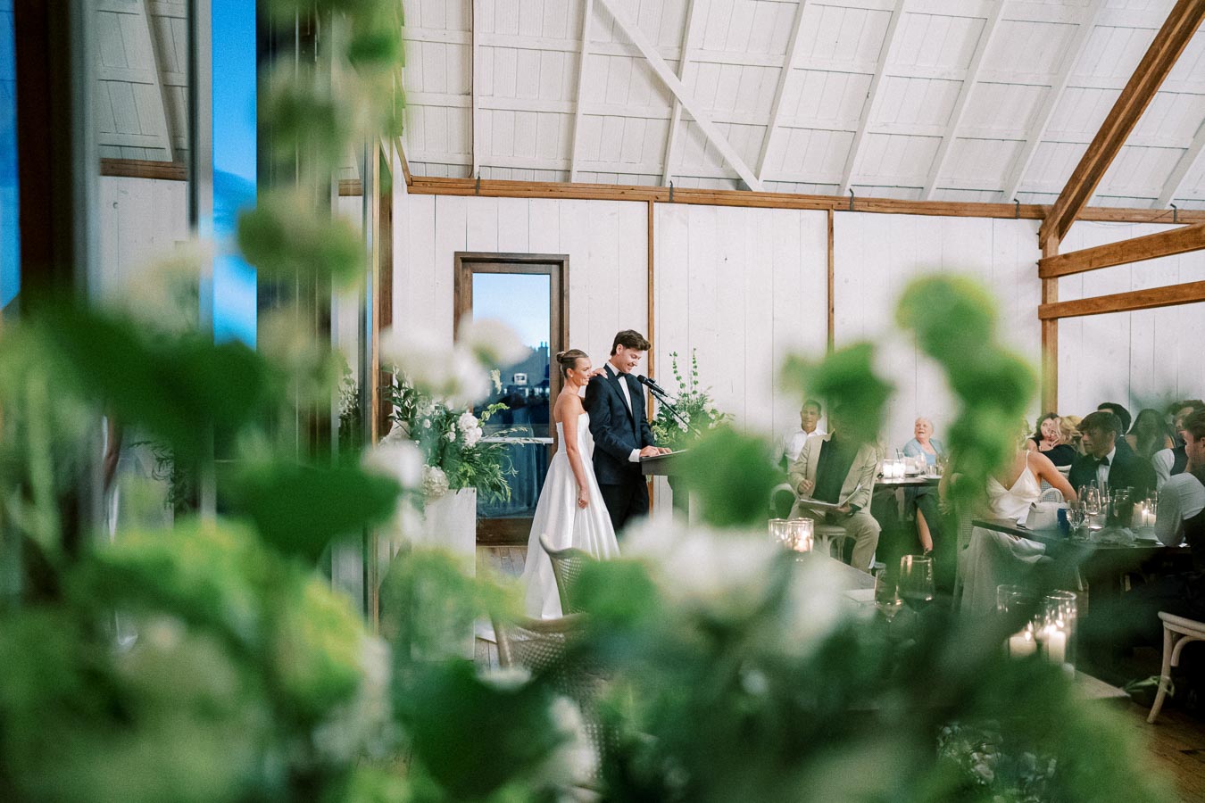 A bride and groom share a moment during their wedding ceremony inside a rustic barn, surrounded by guests seated at decorated tables. The couple stands under a high, white wooden ceiling, framed by lush greenery and white floral arrangements, creating a romantic and elegant ambiance.