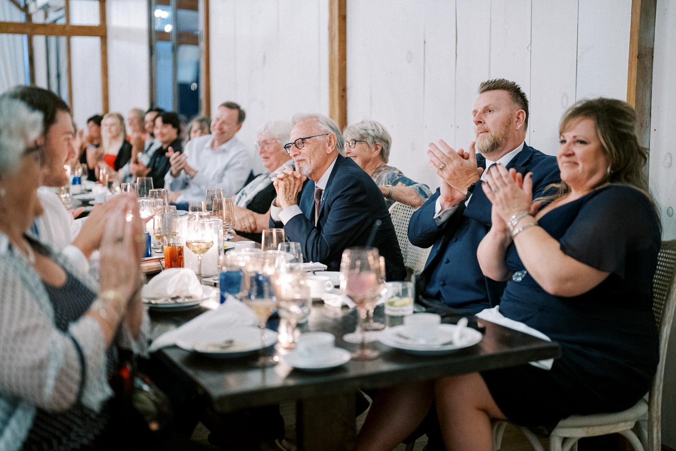 A group of people sitting at a long table dressed in formal attire, clapping and enjoying an event in a cozy, elegantly set dining environment.