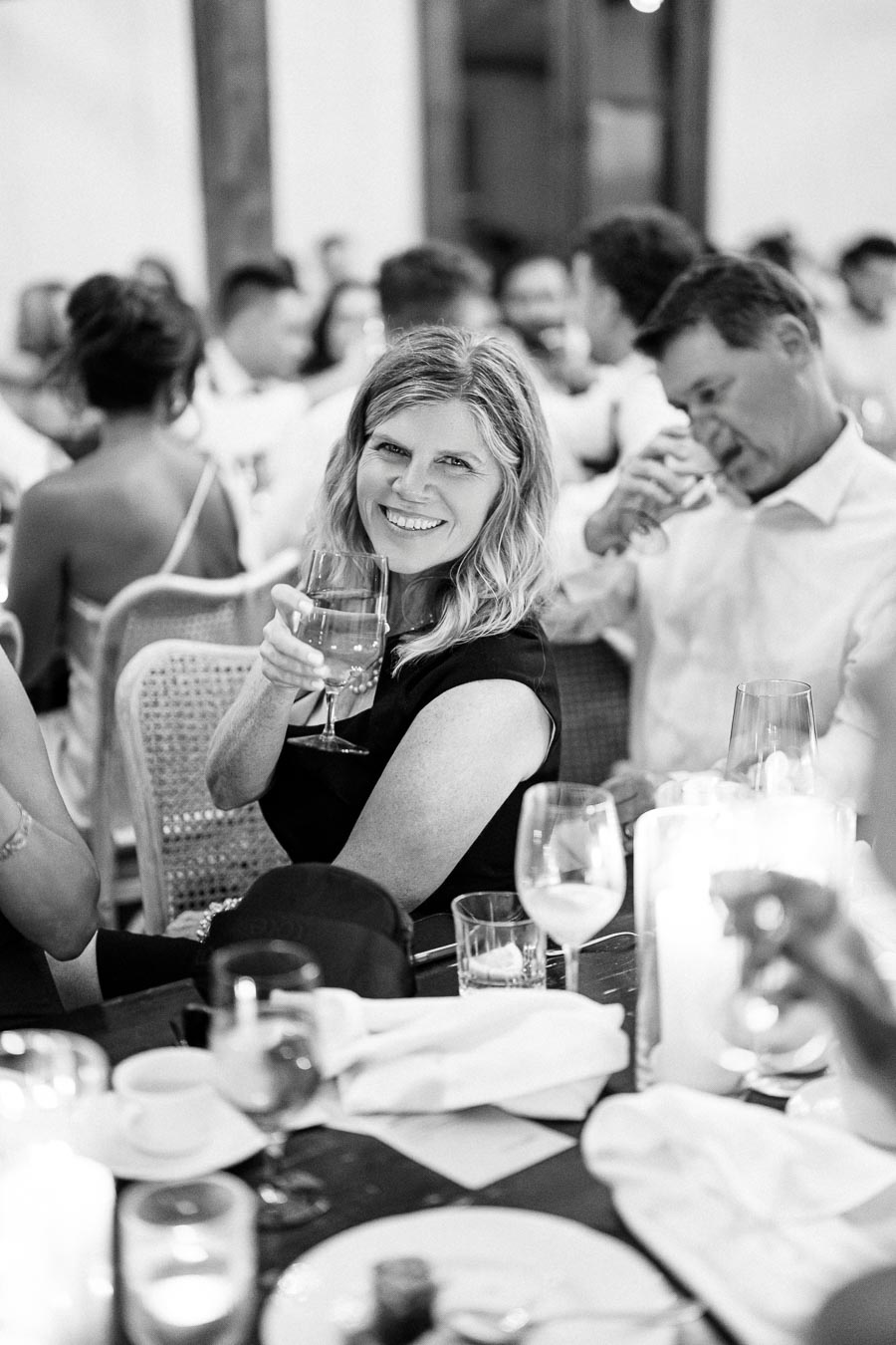 A woman in a black dress smiles and raises a glass at a festive gathering, surrounded by elegantly dressed guests sitting at a table with candles and drinks.