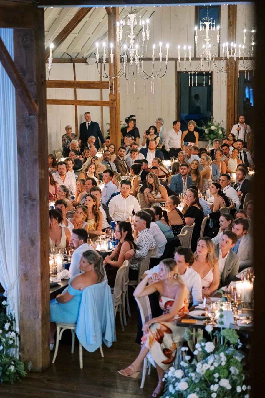 Elegant wedding reception in a rustic barn setting with guests seated at candle-lit tables, dressed in formal attire, and enjoying the evening under ornate chandeliers.
