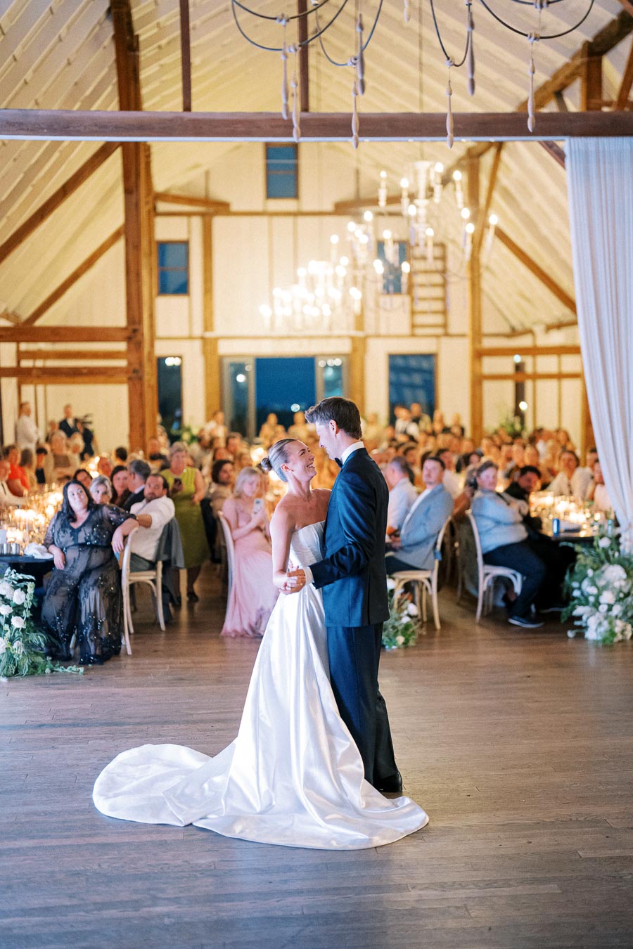 A newlywed couple shares their first dance in an elegant barn wedding venue, surrounded by seated guests and soft candlelight, with wooden beams and chandeliers enhancing the romantic atmosphere.
