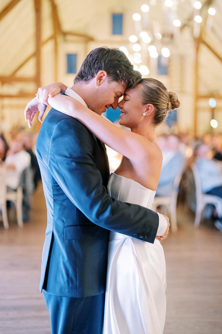 A bride and groom share a romantic first dance at a wedding reception, surrounded by guests in a beautifully lit venue.