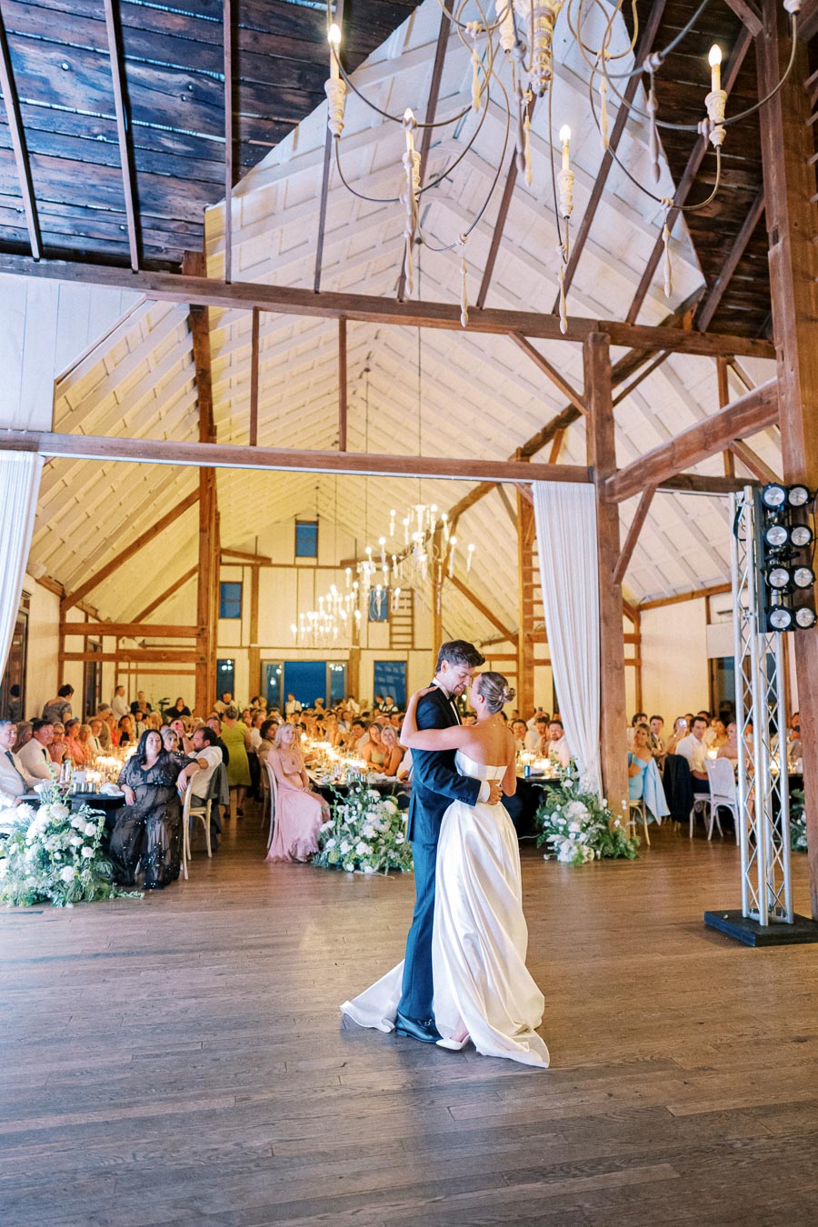A bride and groom share their first dance in a rustic barn wedding venue, surrounded by seated guests and elegant chandeliers, capturing the romantic ambiance of the celebration.