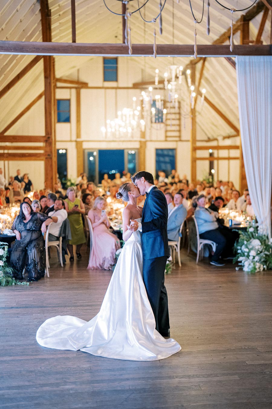 Bride and groom share a kiss during their first dance at a rustic wedding reception with guests seated in a beautifully decorated barn venue.