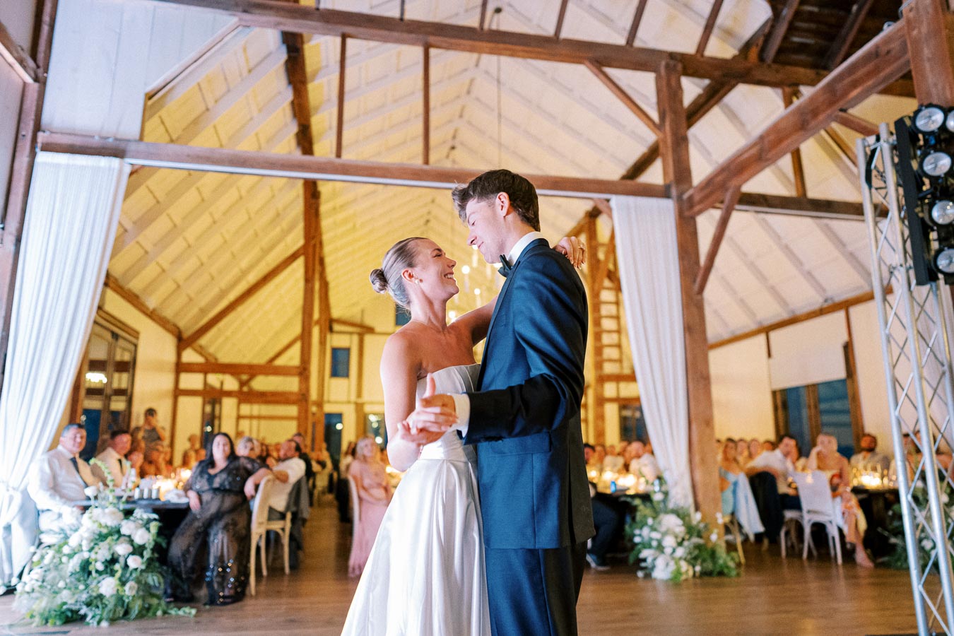 Couple enjoying their first dance at a wedding reception in a rustic barn venue, surrounded by seated guests and elegant floral arrangements.
