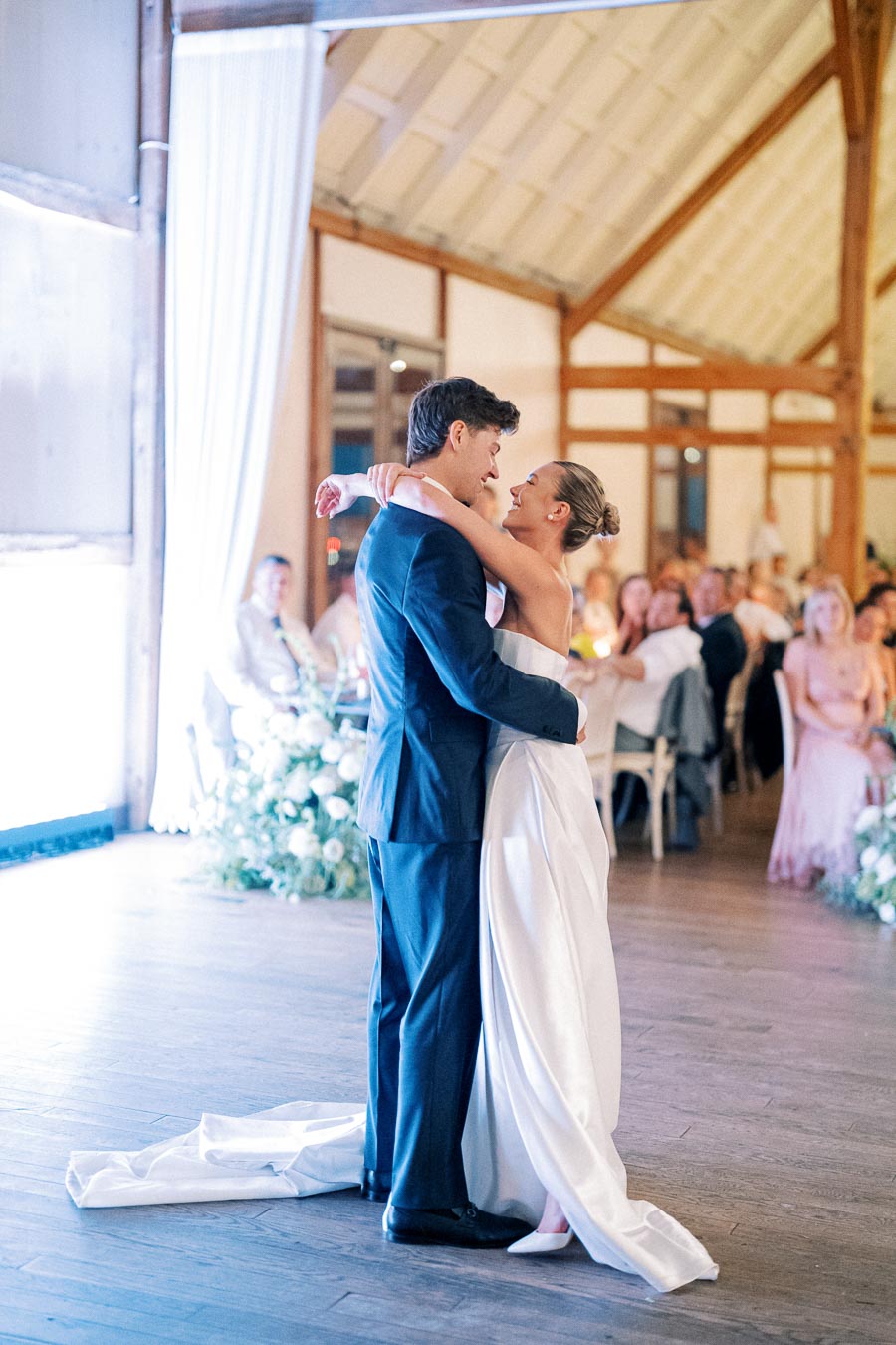 A bride and groom share their first dance at a wedding reception in a warmly lit venue with guests seated in the background.