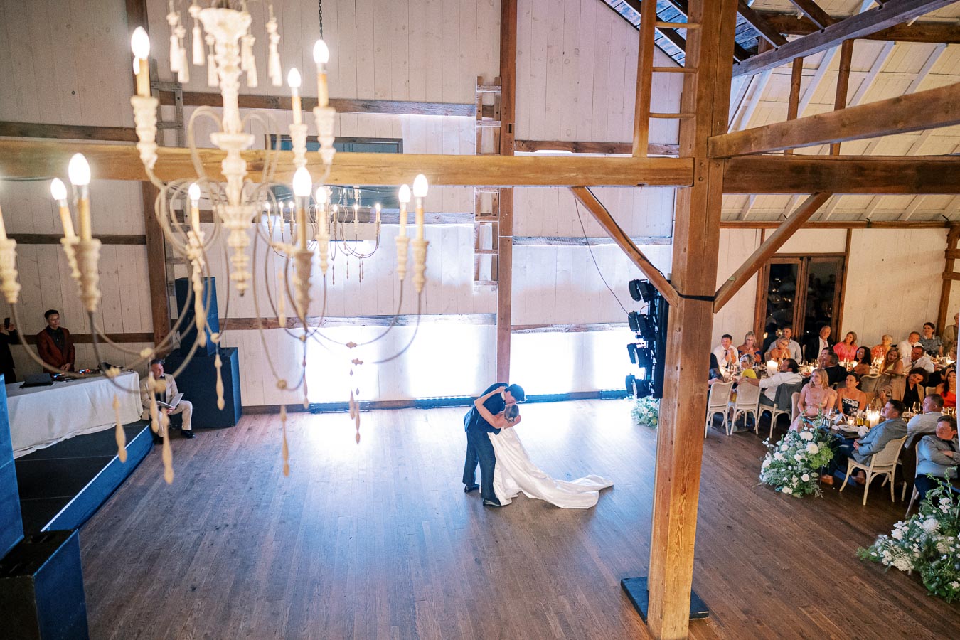 A couple dances in the center of a spacious rustic barn wedding venue under a large white chandelier, surrounded by guests seated at decorated tables.