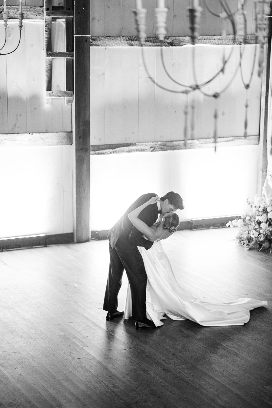 A black and white image of a couple performing a romantic dance in an elegantly decorated wooden hall, with the woman in a flowing gown and the man in a suit gently embracing her in a dance dip.