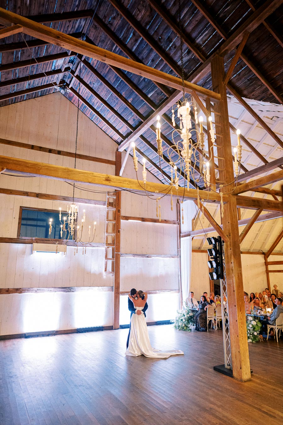 A couple shares a romantic first dance in an elegantly decorated rustic barn with high wooden ceilings and chandeliers, surrounded by seated guests at a wedding reception.