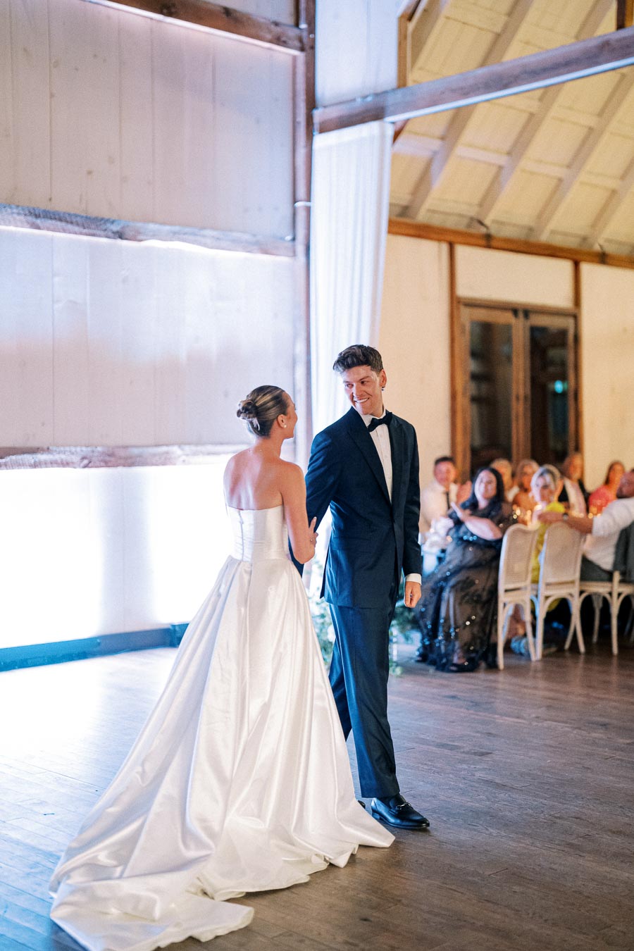 A bride and groom share a moment on the dance floor during their wedding reception in a rustic barn venue, surrounded by guests seated at elegantly decorated tables.