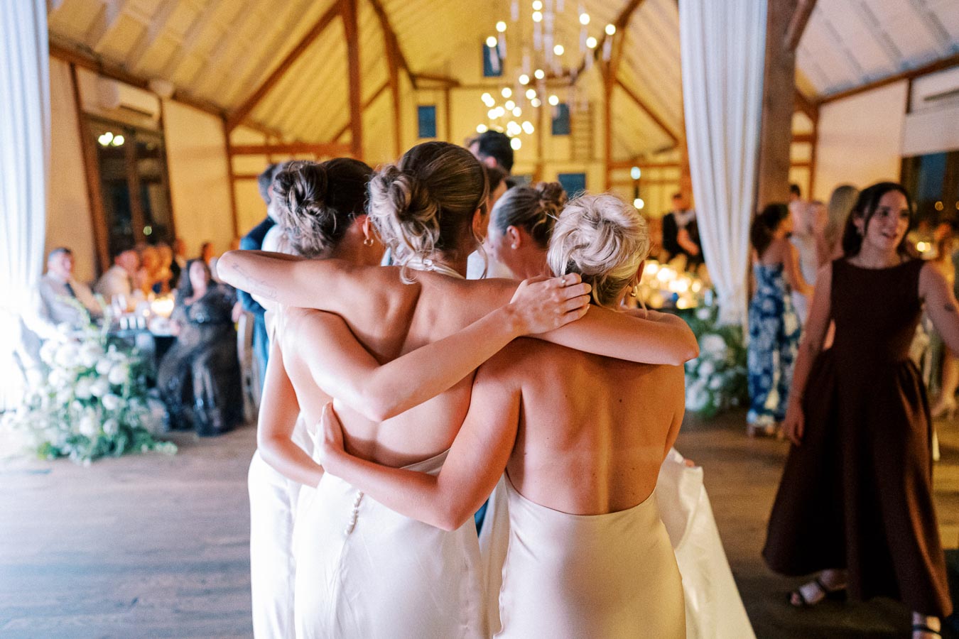 Bridesmaids embracing at a wedding reception in a beautifully lit barn venue.